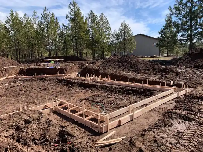 Footing forms staked out in a cleared, muddy lot among pine trees before the pour