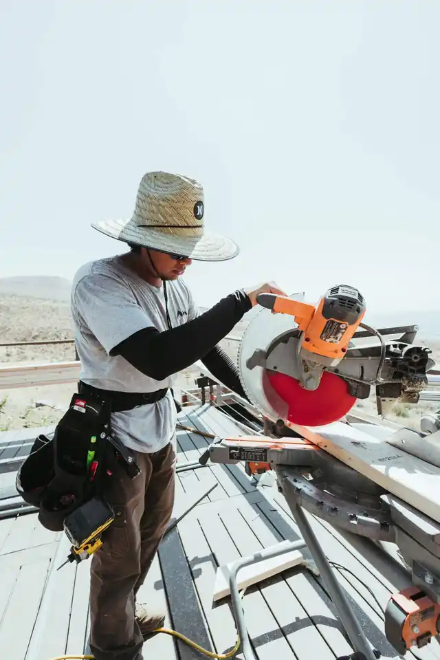 Carpenter in a sun hat cutting lumber on a miter saw at a job site