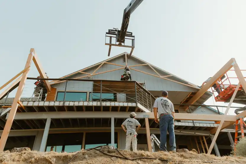 A telehandler lifting a roof truss onto a hillside addition while the crew watches from below